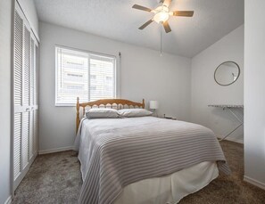 Ein Schlafzimmer mit einem Bett, einem Deckenventilator, einem Fenster mit Jalousien, einem runden Spiegel und einem Bügelbrett.