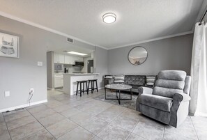 Ein geräumiges Wohnzimmer mit einem grauen Sofa, einem grauen Sessel und einem Couchtisch. Im Hintergrund befindet sich eine Küche mit weißen Schränken und ein Essbereich mit Hockstühlen.