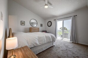 Un dormitorio con cama, lámpara de noche, cómoda, espejo, reloj y puerta corrediza de vidrio a un balcón.