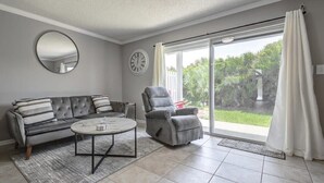 Un salón con sofá, sillón y mesa de centro, un espejo redondo, un reloj y un ventanal corredero que da a un jardín.