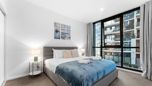 A modern bedroom with a large bed, a nightstand, and a window with a view of a building. - The Amazing City Escape - CBD Modern 1BR Apt (Adelaide)