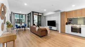 A modern living room with a sofa, a dining table with chairs, a TV, and a kitchen area with a stove and oven. - The Amazing City Escape - CBD Modern 1BR Apt (Adelaide)