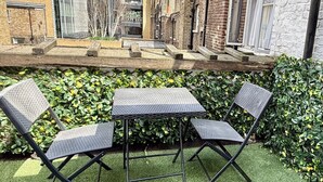 An outdoor seating area with a table and two chairs, surrounded by a hedge and a brick wall.