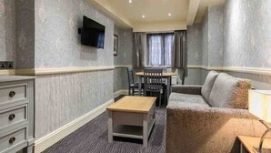 A hotel room with a dining table, chairs, a sofa, a TV mounted on the wall, and a dresser.