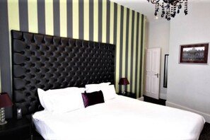 Een slaapkamer met een gestoffeerd hoofdeinde, gestreepte behang, een wit bed en een kroonluchter.