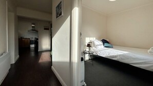 A room with a bed, a bedside table, a lamp, and a small table with a chair. There is a kitchen area visible in the background.