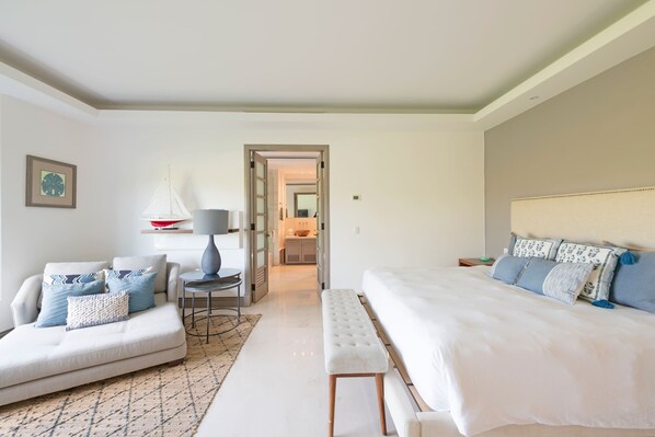 Un dormitorio moderno con una cama grande, un sofá y una banca. Hay una puerta que da a un baño y un pasillo.