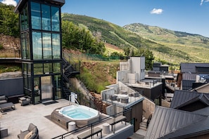 En moderne bygning med et glastårn, udendørs siddepladser, jacuzzi og udsigt over en grøn skråning.