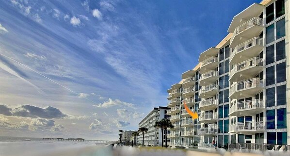 Un hôtel situé en bord de mer, doté de nombreux balcons, avec une jetée en arrière-plan et un ciel dégagé.
