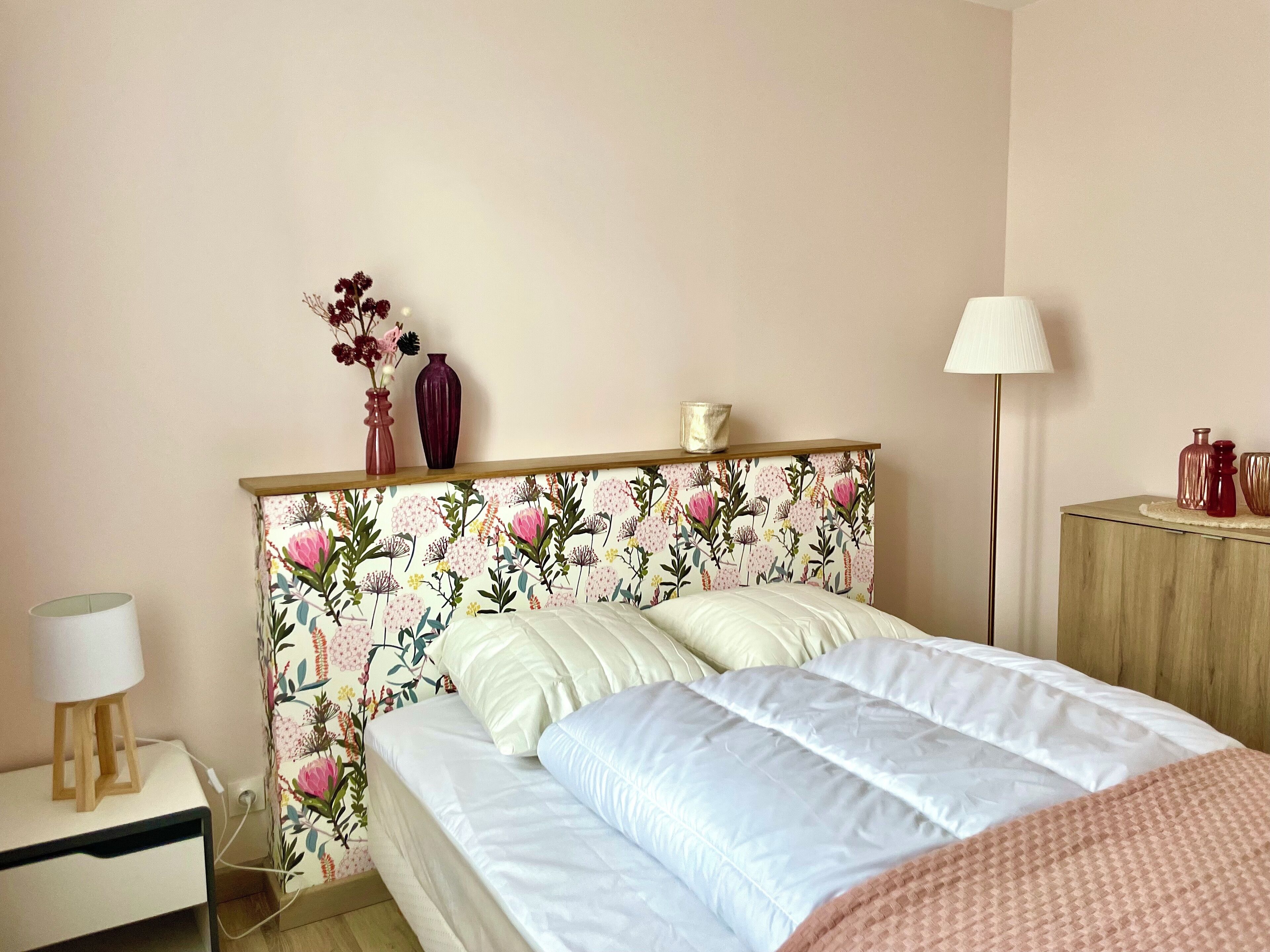 A bedroom with a floral wallpaper headboard, a bed with white bedding, a bedside table with a lamp, and a nightstand with a lamp.