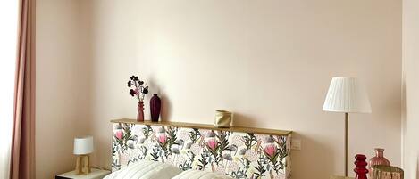 A bedroom with a floral headboard, a bedside table with a lamp, and a bedside plant.