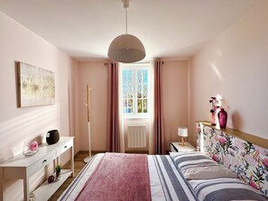 Une chambre à coucher comprenant un lit, des tables de chevet, une lampe sur pied, une fenêtre avec des rideaux et une œuvre d’art accrochée au mur.