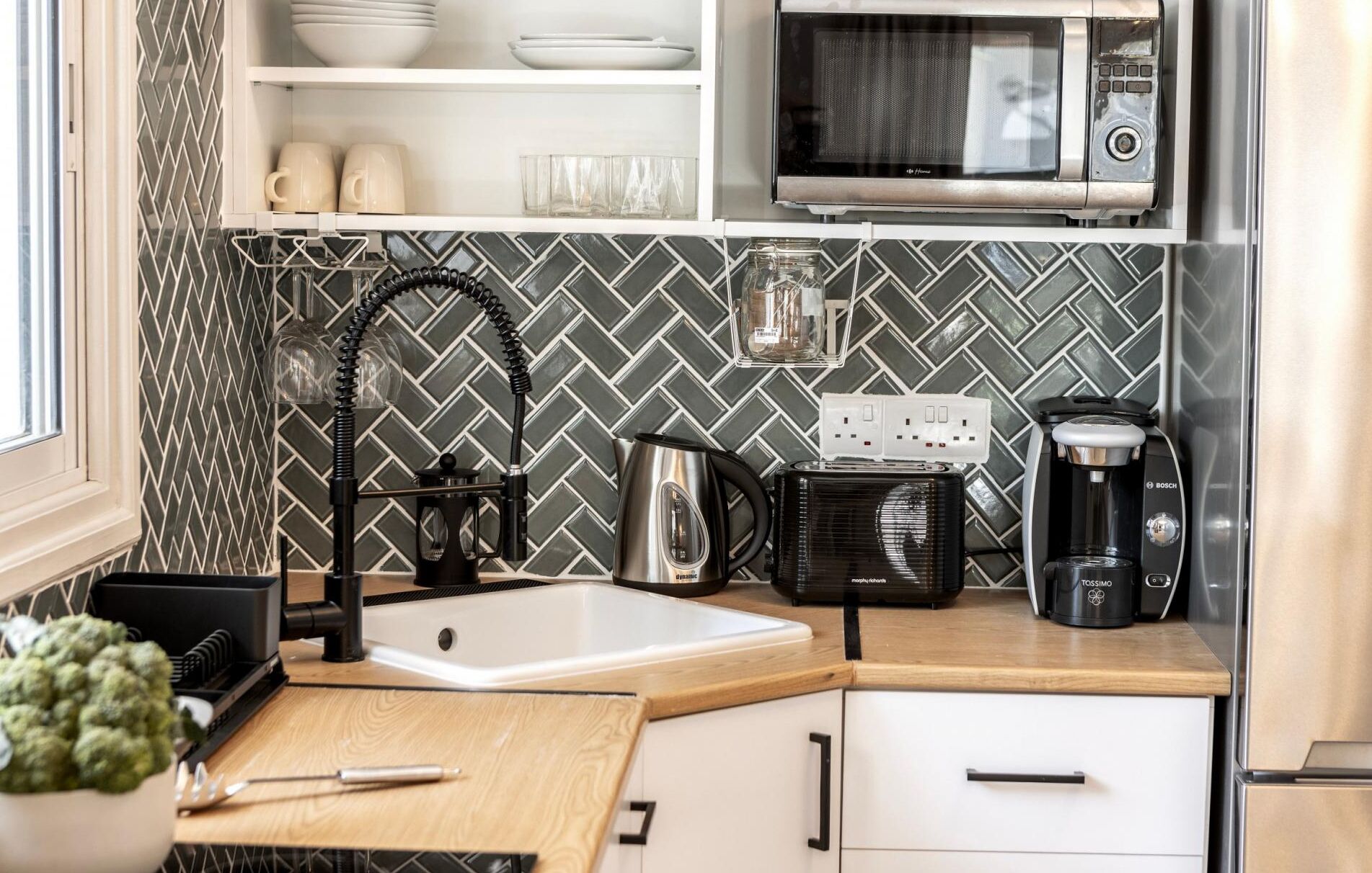 A modern kitchen with a black and white herringbone tile backsplash, a stainless steel kettle, a coffee maker, and a microwave.