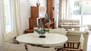 A round dining table with chairs, a ship model, and a television on a wooden stand in a bright living room with a sofa and curtains. - Protaras Apollon Seaside Villa (Paralimni)