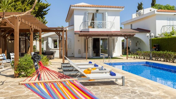 A pool area with lounge chairs, a hammock, and a two-story building with a balcony. - Protaras Apollon Seaside Villa (Paralimni)