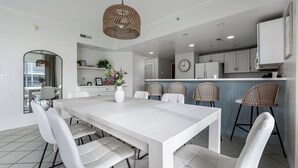 Eine moderne Küche mit weißen Schränken, ein Essbereich mit weißem Tisch und Stühlen, und Weidekörbchenstühle im Hintergrund.