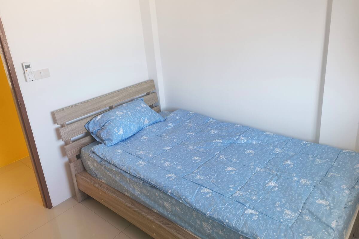 Une petite chambre avec un lit simple, une tête de lit en bois, une couvre-lit à motifs floraux bleus et un mur blanc uni.