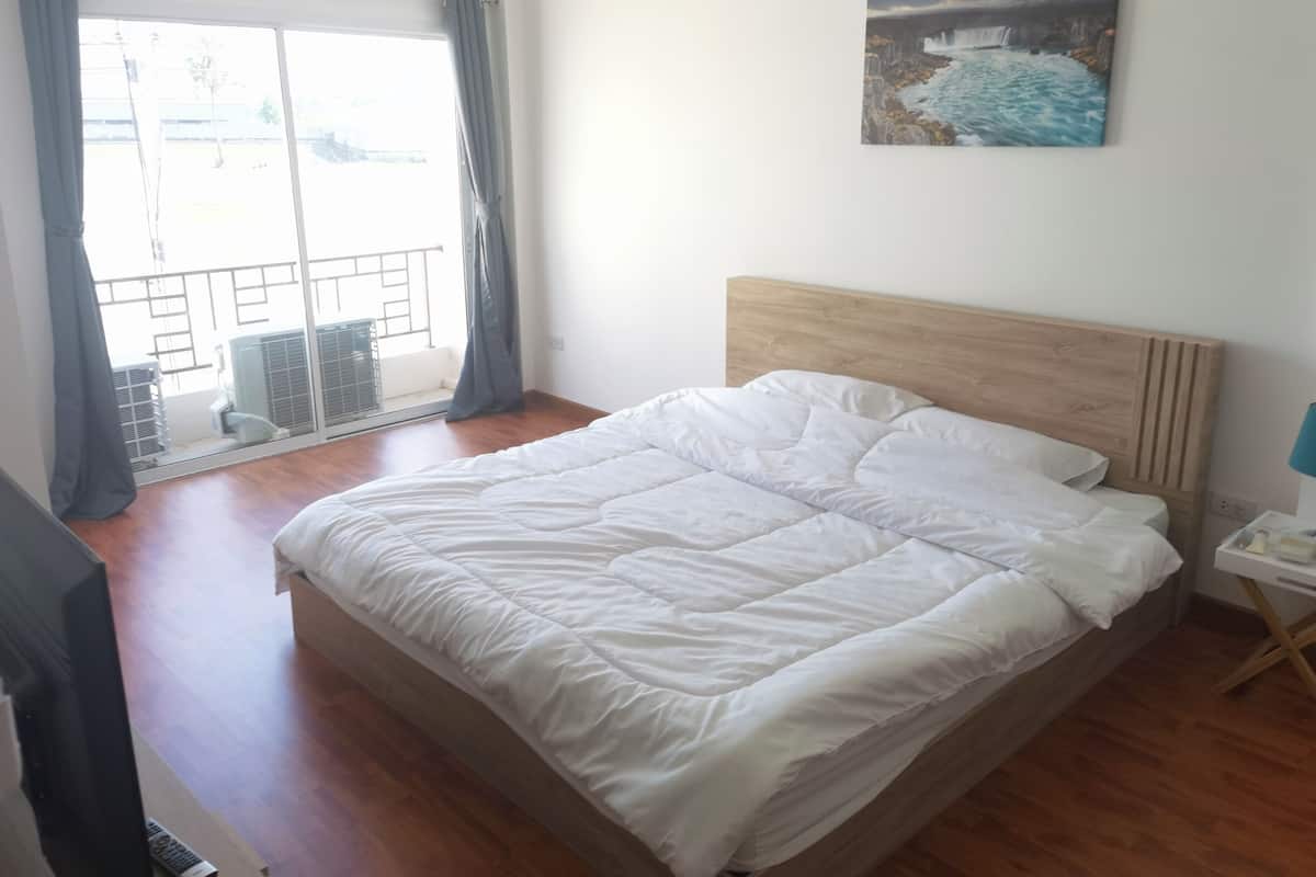 Une chambre à coucher avec une tête de lit en bois, un grand lit avec une literie blanche, un tableau accroché au mur et une vue sur l’extérieur à travers une fenêtre ornée de rideaux.