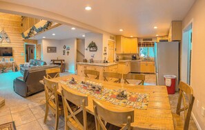 Une cuisine chaleureuse avec des meubles en bois, une table à manger recouverte d’une nappe colorée et un coin salon doté d’un canapé en cuir et d’un téléviseur.