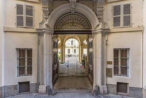 Une entrée imposante, avec un portail en arc de cercle, flanquée de colonnes et d’éléments décoratifs en métal, menant à un bâtiment doté de nombreuses fenêtres et d’une entrée fermée par un portail.