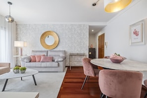 Een moderne woonkamer met een grijze bank, een ronde spiegel, een eettafel met roze stoelen en een klein bijzettafeltje met een lamp.
