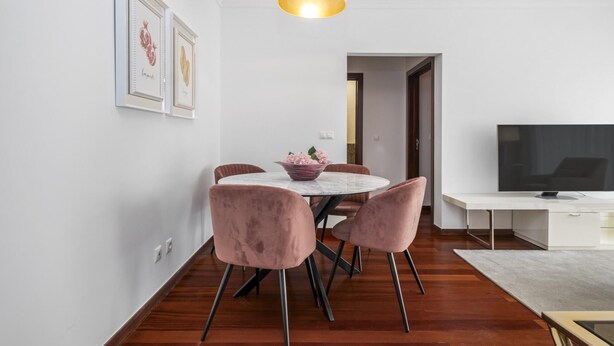 Un área de comedor moderna con una mesa redonda, sillas rosa, una lámpara de techo blanca y un televisor de pantalla plana sobre un mueble blanco.