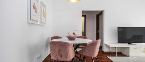 Un área de comedor moderna con una mesa redonda, sillas rosa, una lámpara de techo blanca y un televisor de pantalla plana sobre un mueble blanco.