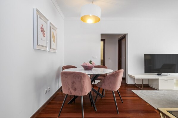 Een moderne eetruimte met een ronde tafel, roze stoelen, een witte plafondlamp en een flatscreen televisie op een witte kast.