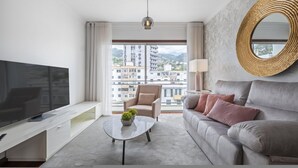Una habitación de hotel moderna con una gran televisión de pantalla plana, un sofá, una mesa de centro con una planta, una silla y un ventanal con vistas a la ciudad.