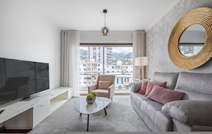 Een moderne hotelkamer met een grote flatscreen-tv, een bank, een bijzettafeltje met een plant, een stoel en een groot raam met uitzicht op de stad.