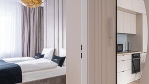 A modern bedroom with a large bed, a hanging light fixture, and a view into the adjacent kitchen area. - RentPlanet - Nowa Manufaktura (Wroclaw)