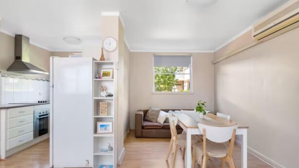 Et kompakt køkken med hvide skabe, en indbygget ovn og et hvidt køleskab. Der er et spiseområde med et hvidt bord og stole, samt en brun sofa.