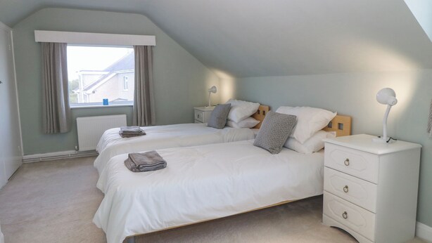 A bedroom with two beds, a nightstand, a dresser, and a window with curtains. - Awelfryn (Holyhead)
