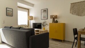 En moderne stue med en grå sofa, et fjernsyn, et gult skab og et spiseområde med bord og stol.
