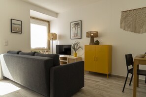 A modern living room with a grey sofa, a television, a yellow cabinet, and a dining area with a table and chair.