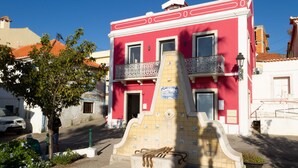 Un edificio rojo con un balcón, un monumento con azulejos en frente y un árbol a la izquierda.
