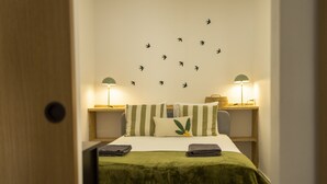 Una cama bien hecha con una manta verde, dos almohadas y un adhesivo mural de pájaros. Hay dos lámparas de noche de madera.