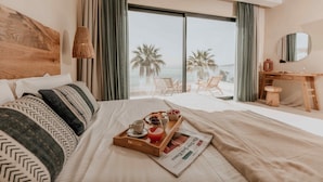 A hotel room with a bed, a wooden headboard, a desk with a chair, a window with a view of palm trees, and a tray with breakfast items. - Ostrako Beachside Suites (Naxos)
