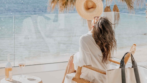 A person sitting by a glass table with a bottle and glass, overlooking a beach with palm trees and the sea. - Ostrako Beachside Suites (Naxos)