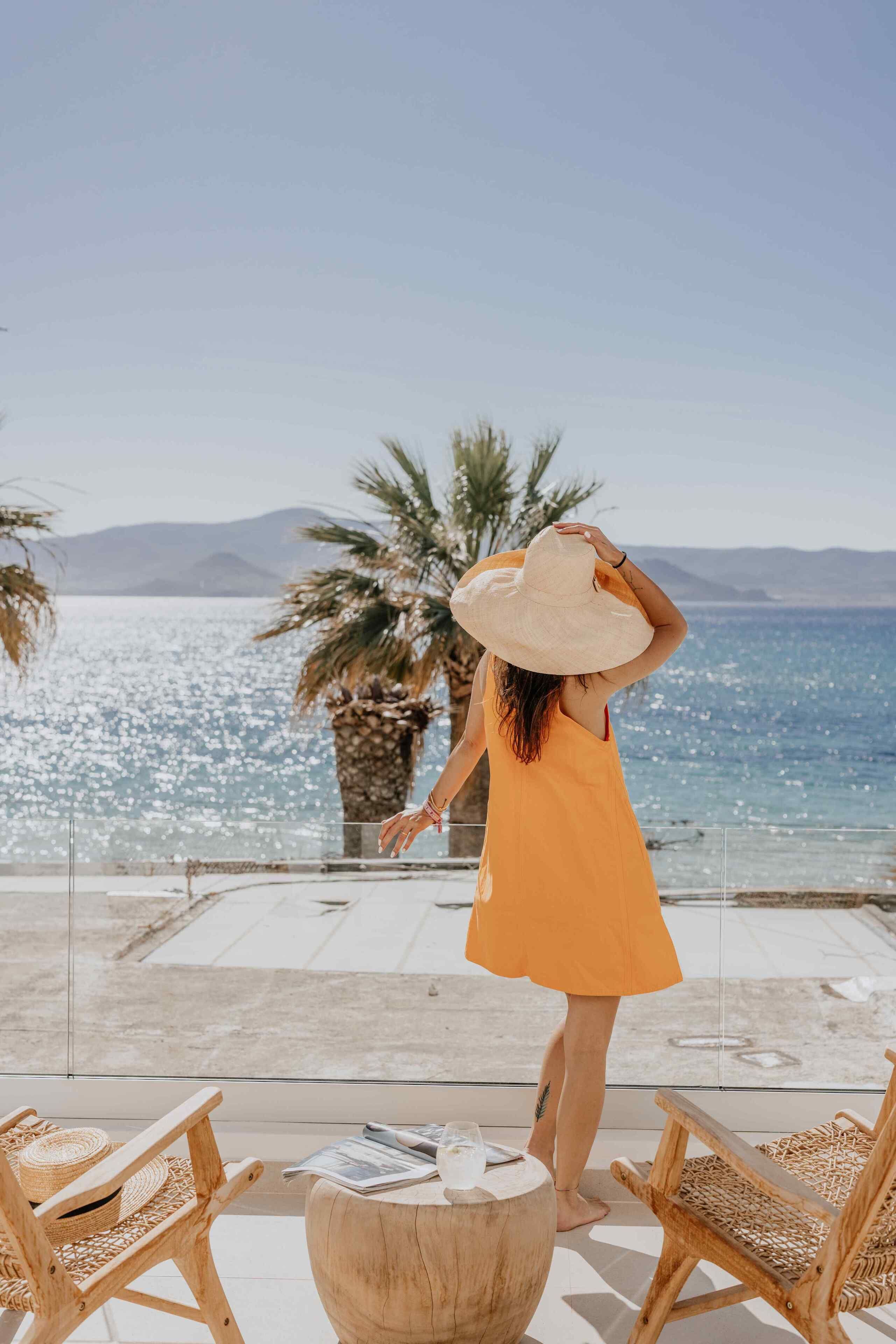 A person in an orange dress standing by a seaside with a large hat, overlooking a beach with palm trees and a clear sky.