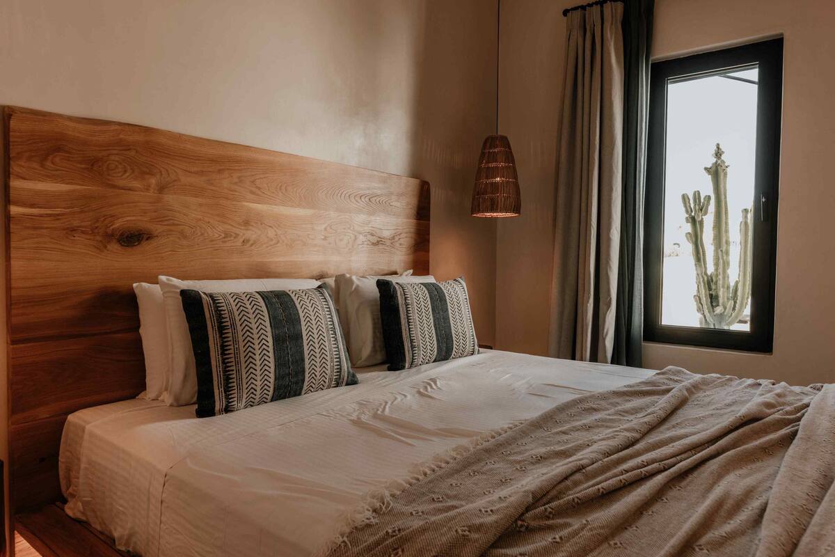 A bedroom with a wooden headboard, a bed with patterned pillows, a hanging light fixture, and a window with a cactus view.