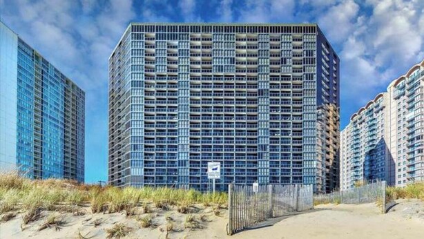 A high-rise building with a grid of windows, surrounded by a sandy area with sparse vegetation. - Direct Ocean Front Studio at the Golden Sands with Indoor Pool, Gym and Saunas (Ocean City)