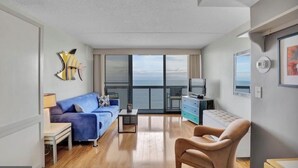 A modern living room with a blue sofa, a beige armchair, a coffee table, and a TV. There is a large window with a view of the sea. - Direct Ocean Front Studio at the Golden Sands with Indoor Pool, Gym and Saunas (Ocean City)