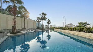 Piscina com espreguiçadeiras e palmeiras, um edifício moderno e uma roda-gigante ao fundo.