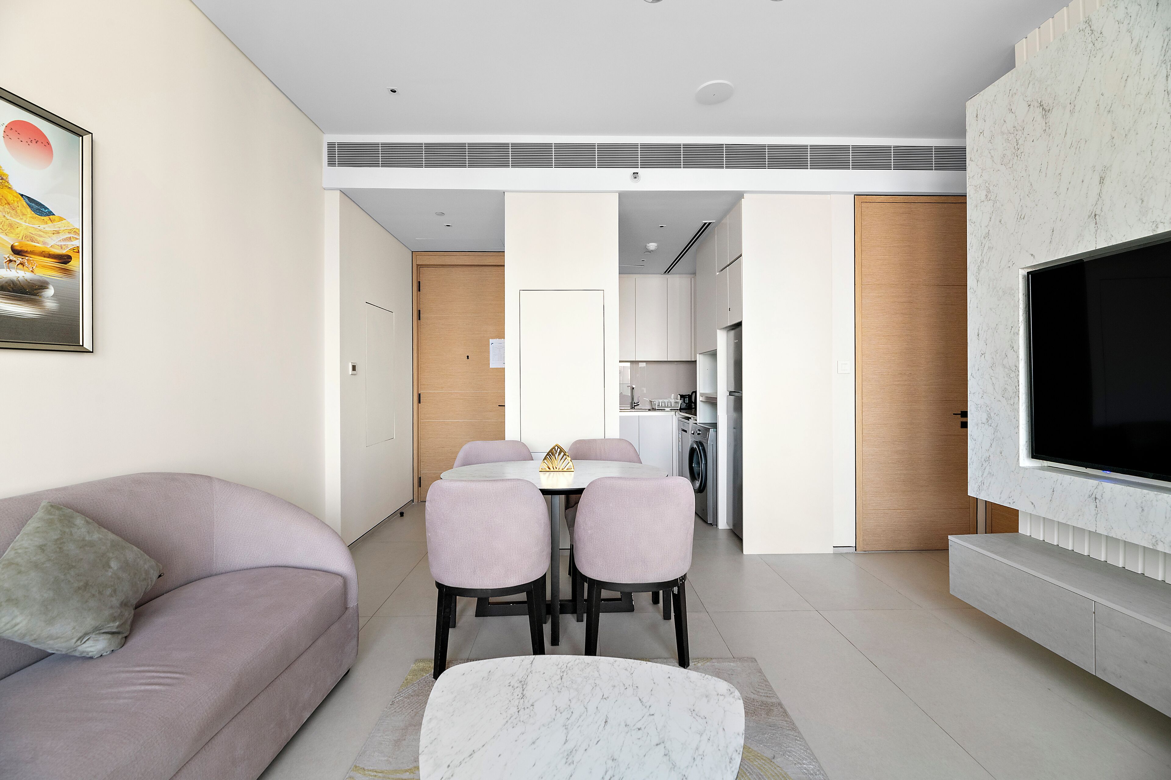 A modern living room with a sofa, a dining table with chairs, a TV mounted on the wall, and a kitchen in the background.