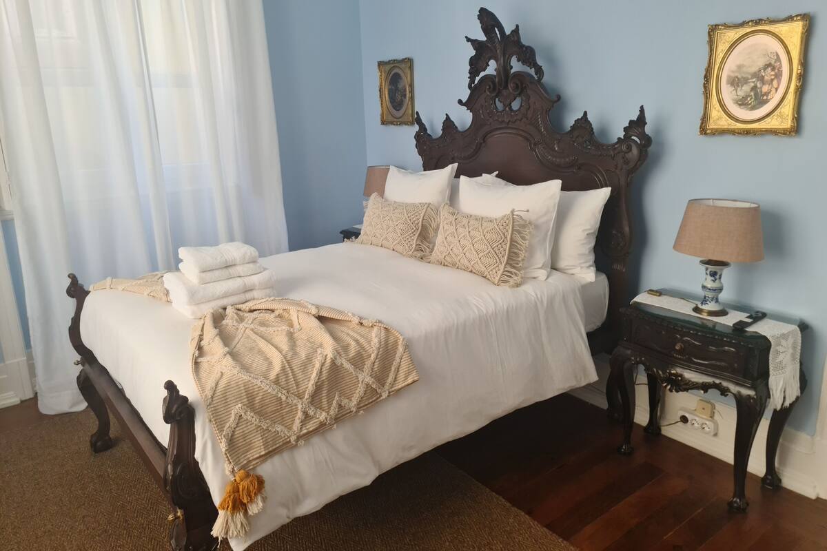 Un lit bien fait, avec du linge de lit blanc et une tête de lit décorative, une table de chevet avec une lampe, et deux tableaux encadrés accrochés au mur.