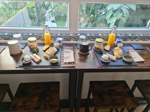Deux tables de petit-déjeuner, dressées devant une fenêtre donnant sur un paysage verdoyant, et sur lesquelles sont disposés divers aliments et boissons.