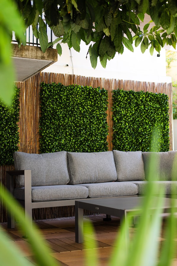 A modern outdoor seating area with a grey sofa and a table, surrounded by a bamboo fence and lush green foliage.