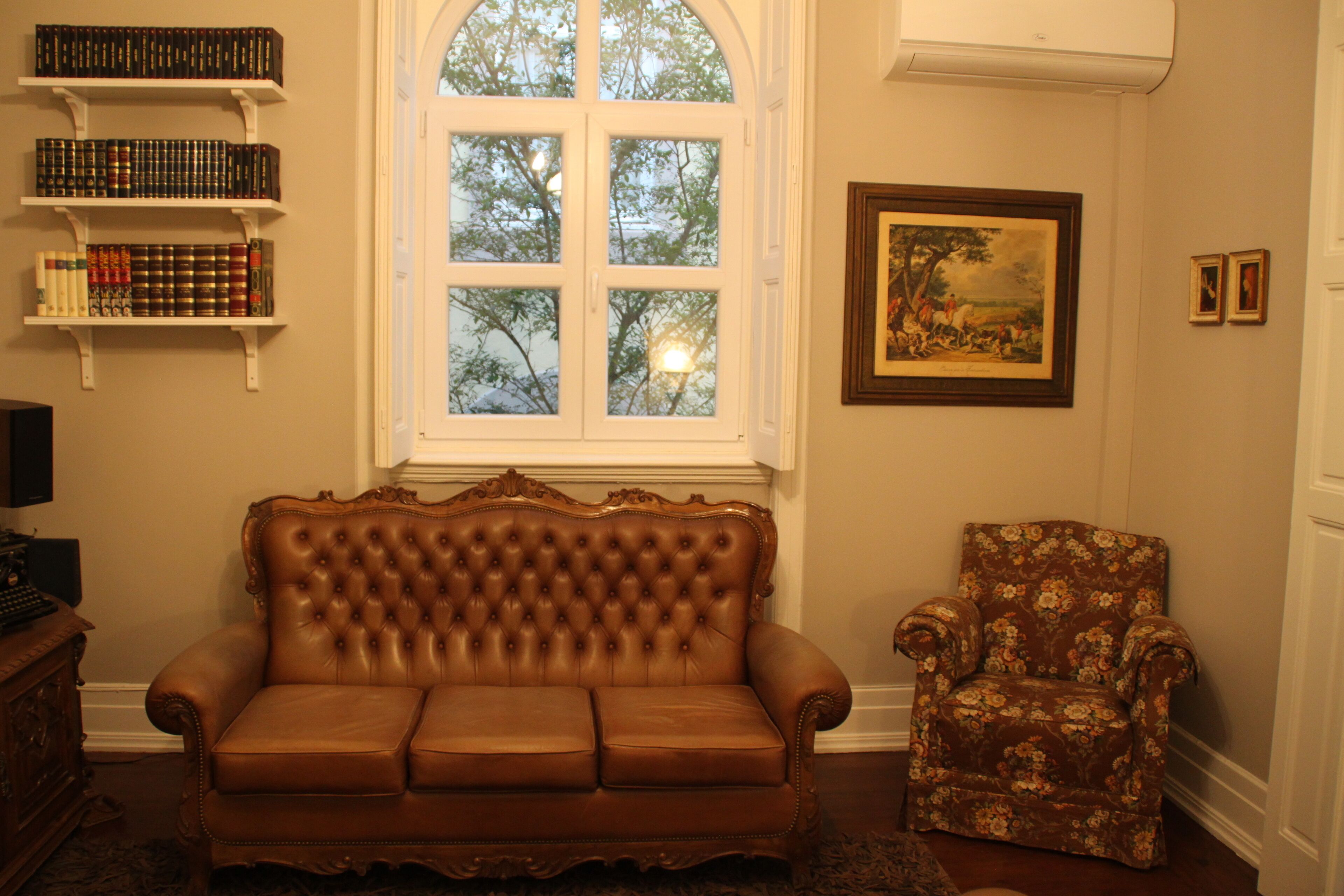 A room with a brown leather sofa, a floral armchair, a bookshelf, a framed painting, and a window with a view of trees.