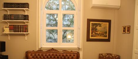 A room with a brown leather sofa, a floral armchair, a bookshelf, a framed painting, and a window with a view of trees.